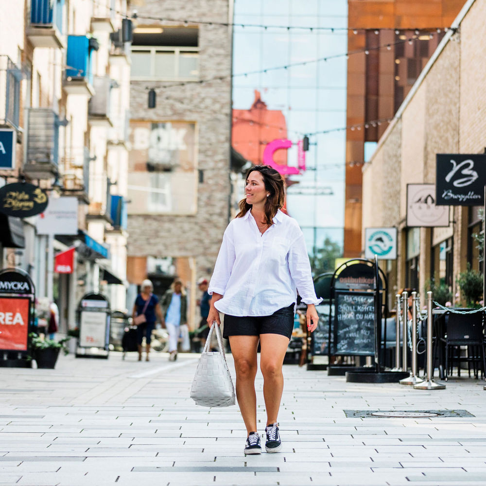 Mörkhårig ung kvinna iklädd vit skjorta, svarta shorts och sneakers bär på en vit väska på promenad i Mölndals innerstad. En gata med butiker på sidorna. 