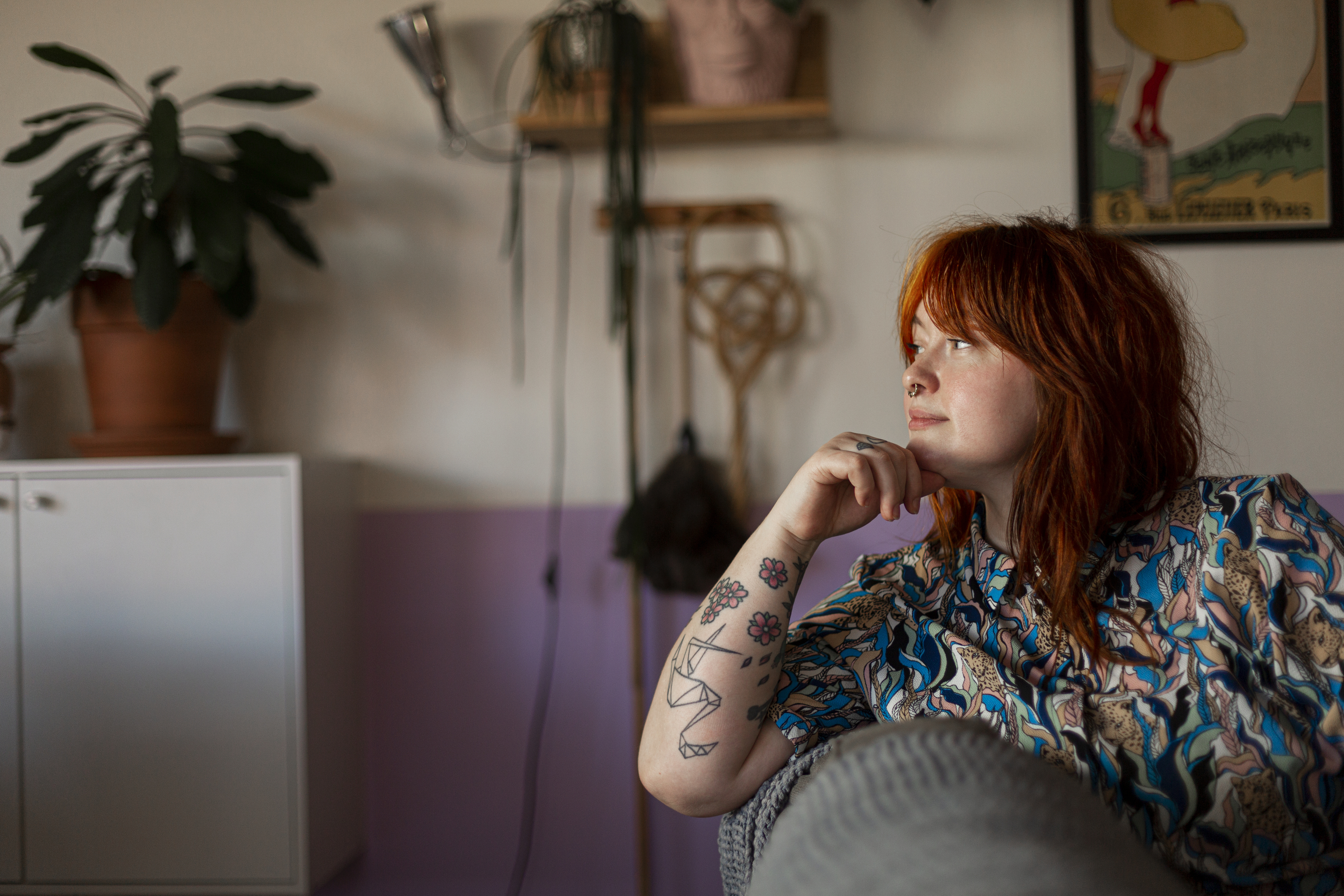 Närbild på ung rödhårig kvinna funderar sittandes i sin soffa, i bakgrunden syns diverse städredskap hängandes på väggen. 