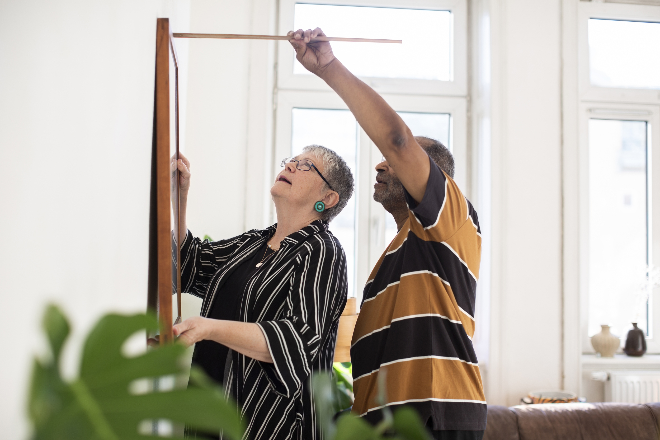 En medelålders man i randig t-shirt mäter på en vägg ovanför en tavla som en kvinna i kort grått hår och glasögon står och håller i. De ska ska sätta upp en tavla i lägenheten som är vit med vita fönster i bakgrunden. 