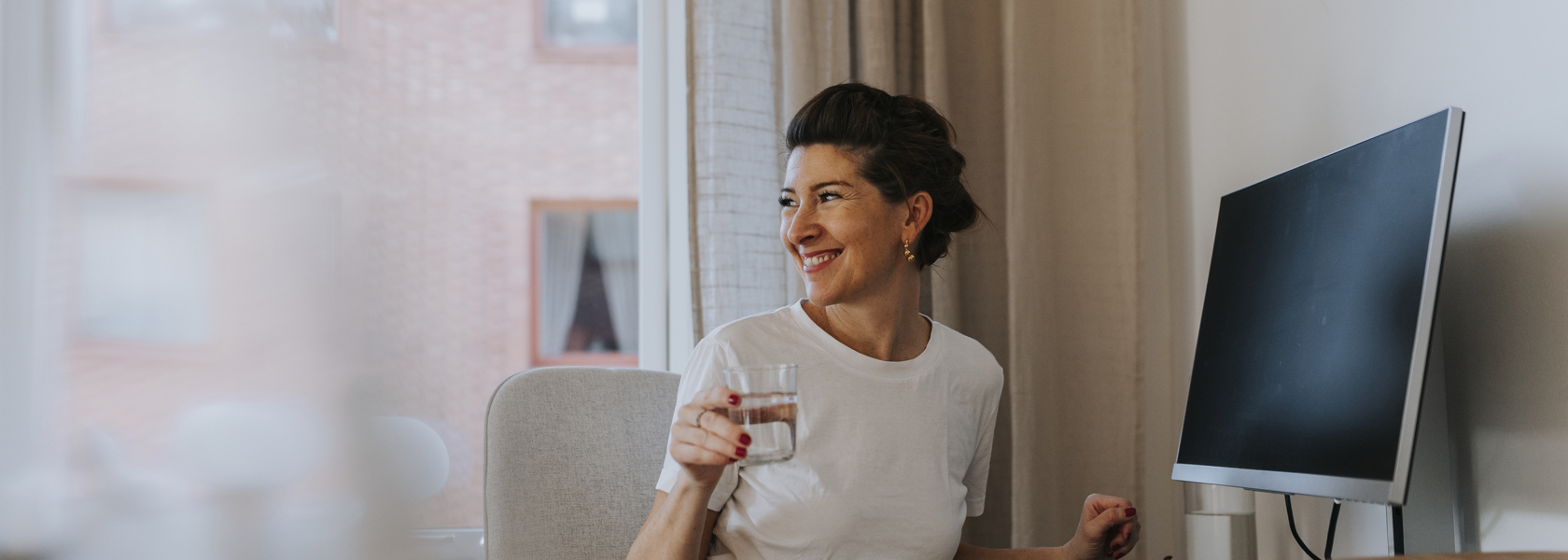 Mörkhårig leende kvinna iklädd vit t-shirt med uppsatt hår sitter bortvänd från en dator i ett hem med ett glas i handen. 