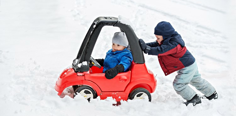ett litet barn skjuter på ett annat barn som sitter i en liten leksaksbil omringade av mycket snö. 