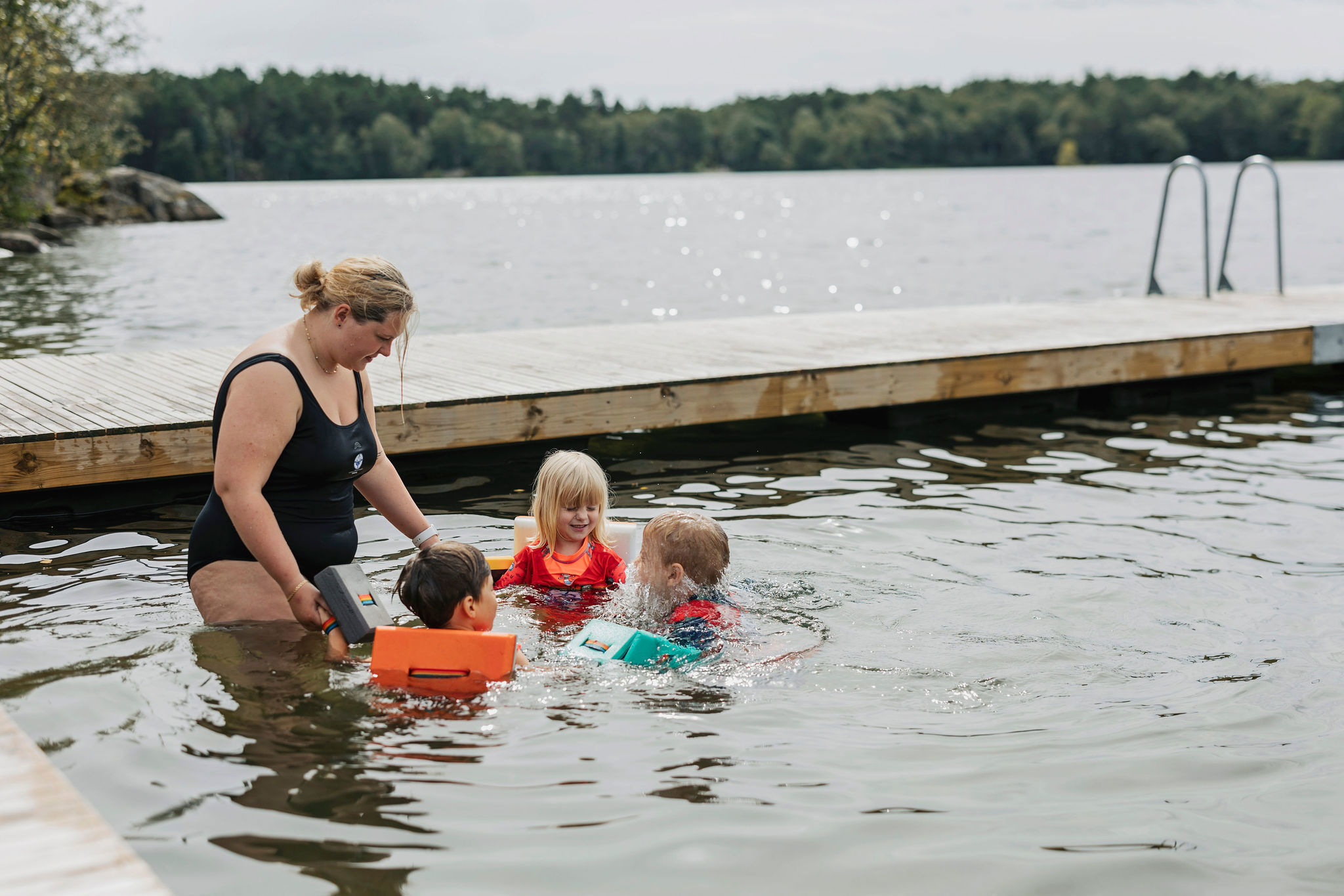 En simlärare och tre barn står i en ring och håller varandra i handen vid en brygga i vattnet. 