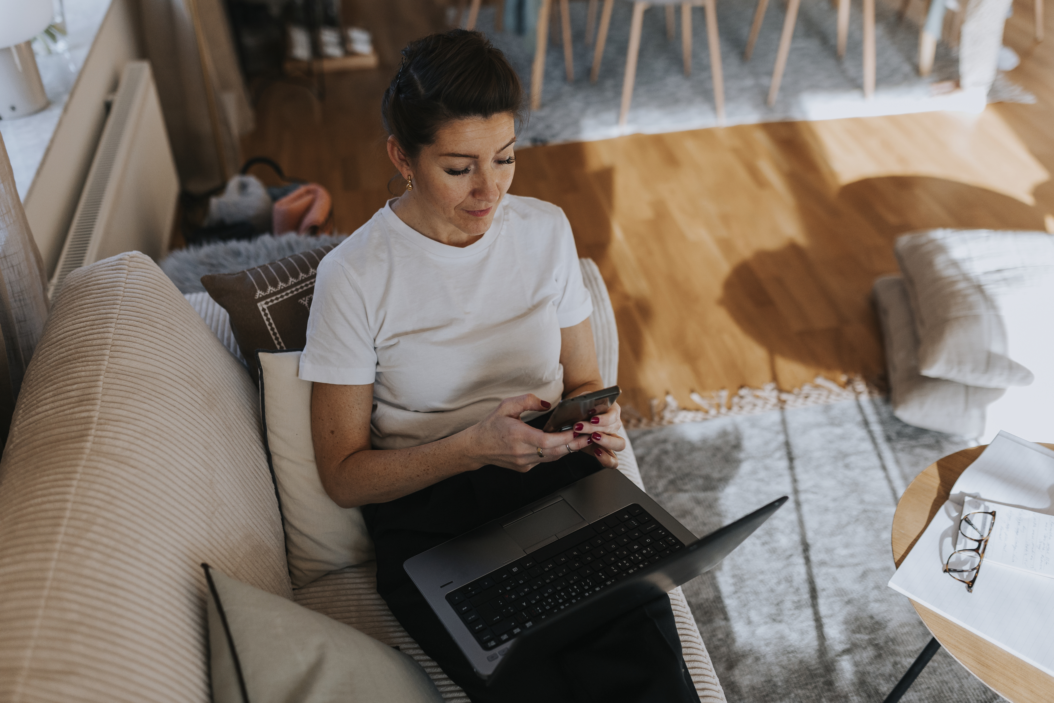 Kvinna i en soffa tittar på sin mobiltelefon och har en laptopdator i knät.