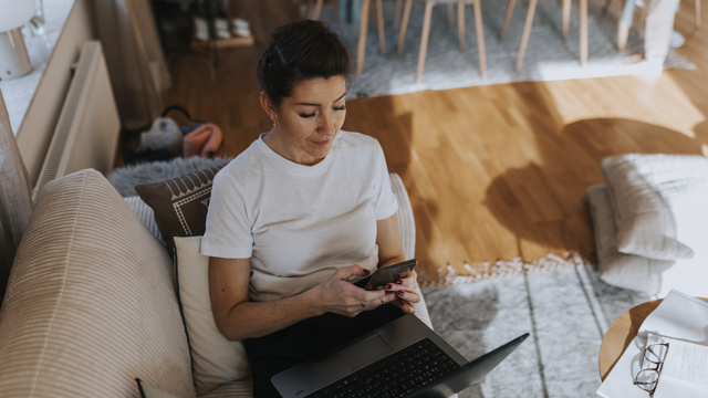 Kvinna i en soffa tittar på sin mobiltelefon och har en laptopdator i knät.