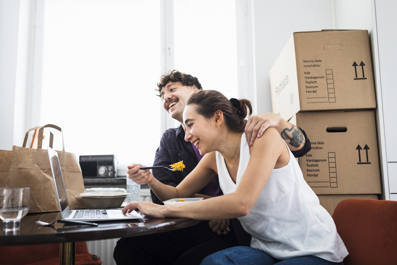 Ungt par skrattar framför en laptop omgiven av flyttlådor.