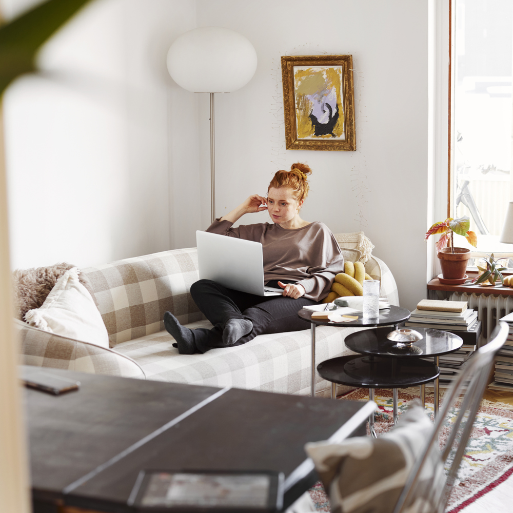 Ung kvinna sitter i sin soffa med en dator i knät i ett ljust vardagsrum. 