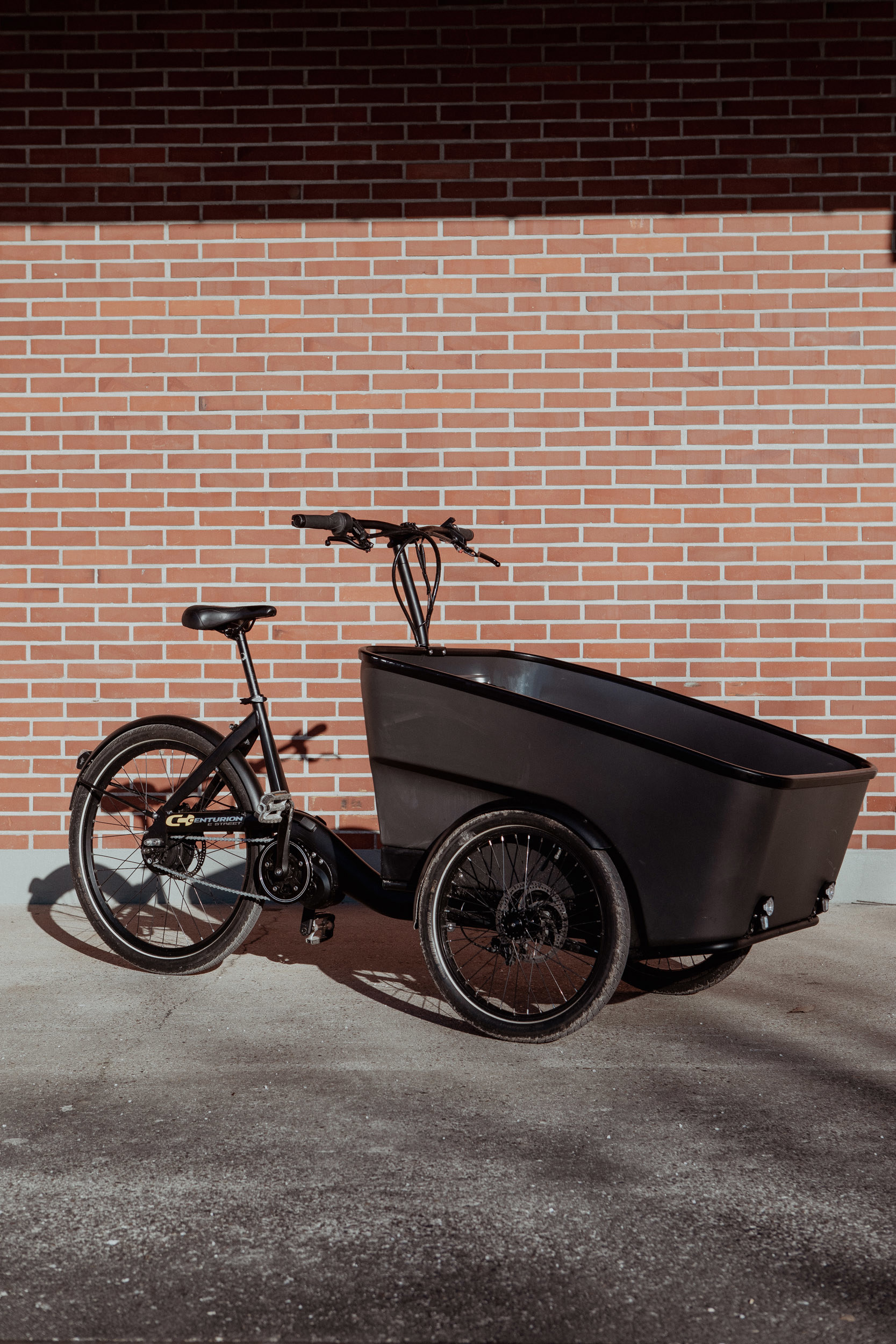 En svart lådlastcykel parkerad framför en röd tegelvägg.