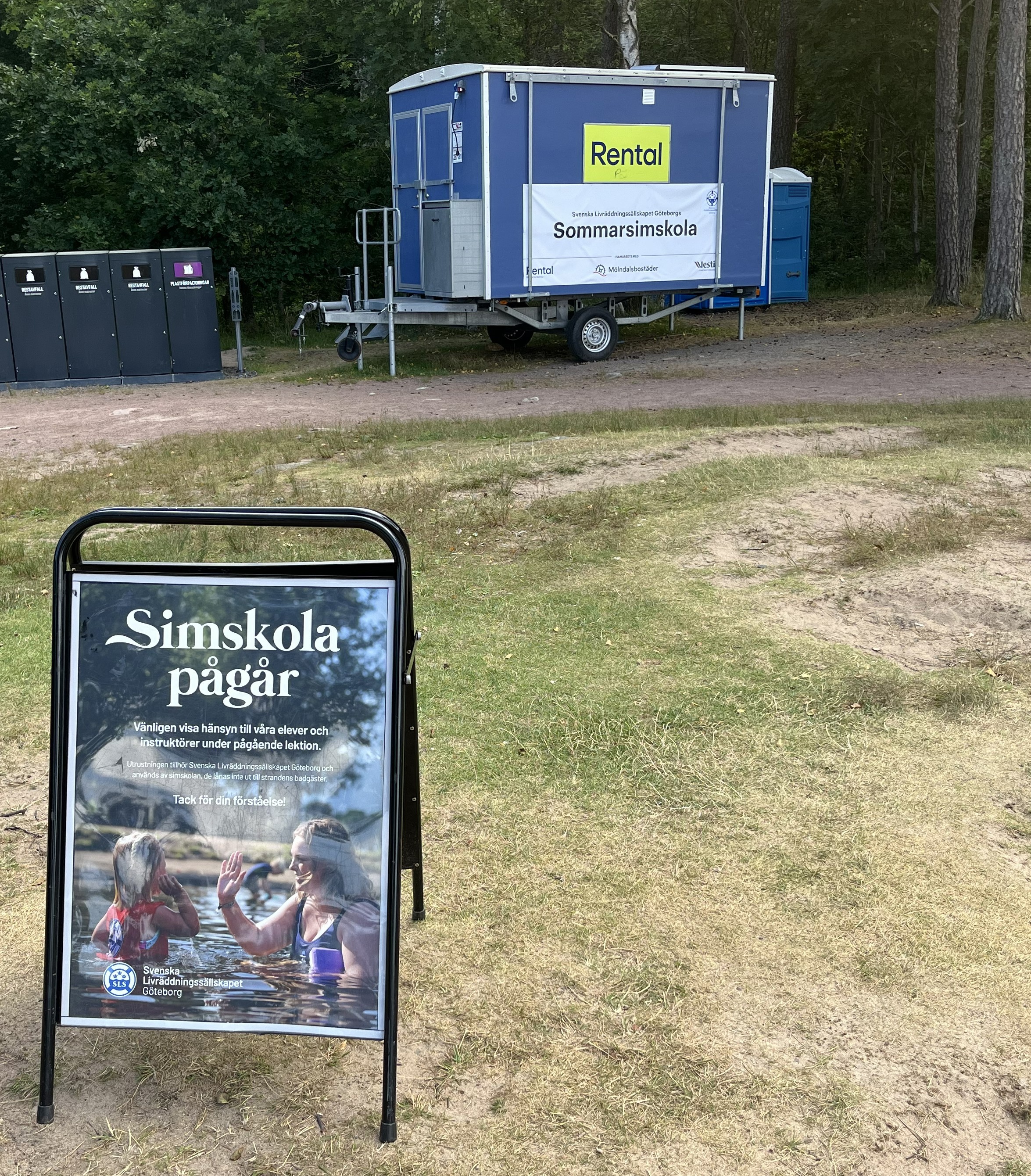 En skylt i förgrunden där det står simskola pågår och en bod mot en skog. 