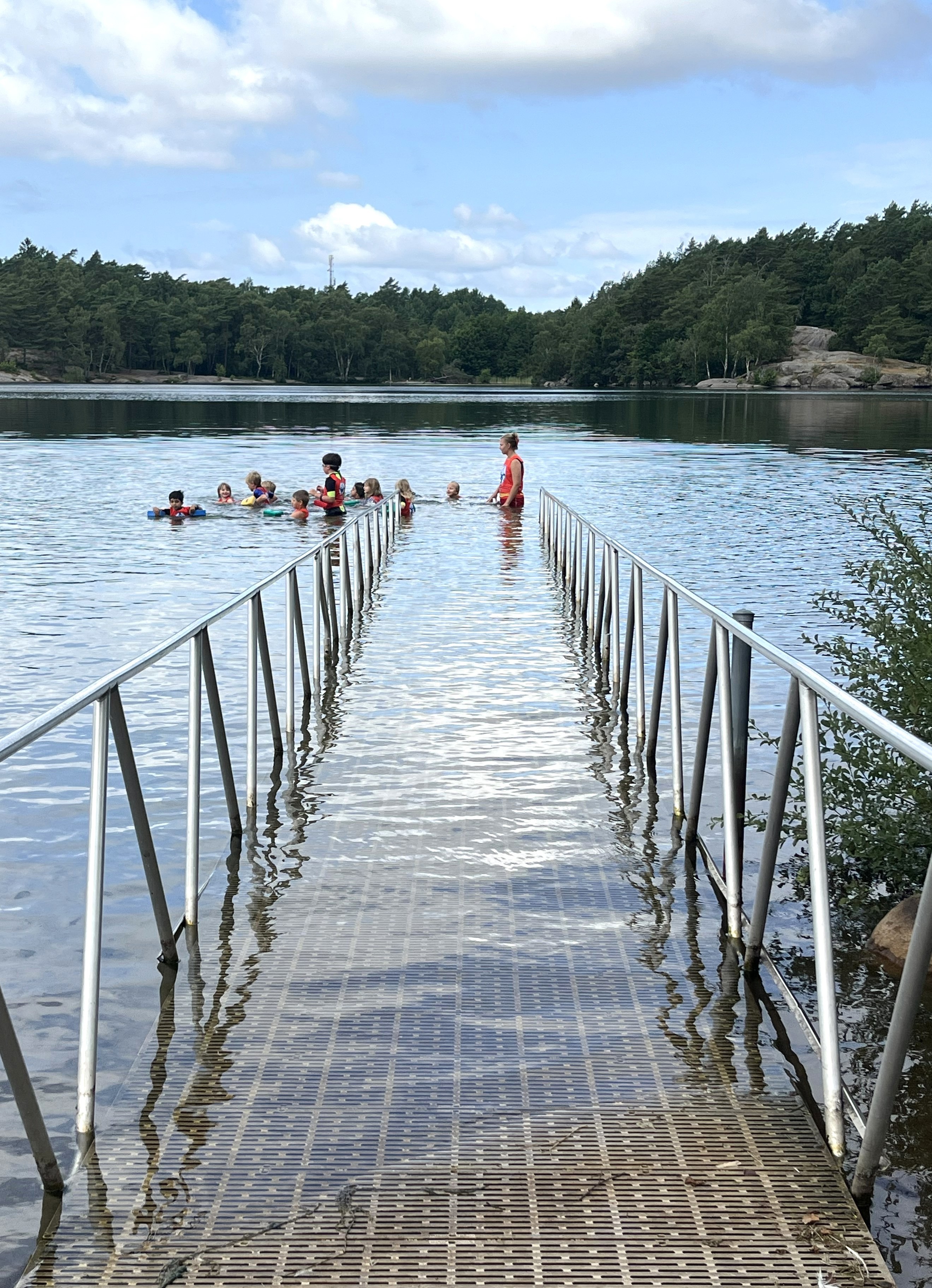 En ramp leder ner i sjön där en grupp barn står tillsammans med simskoleläraren.